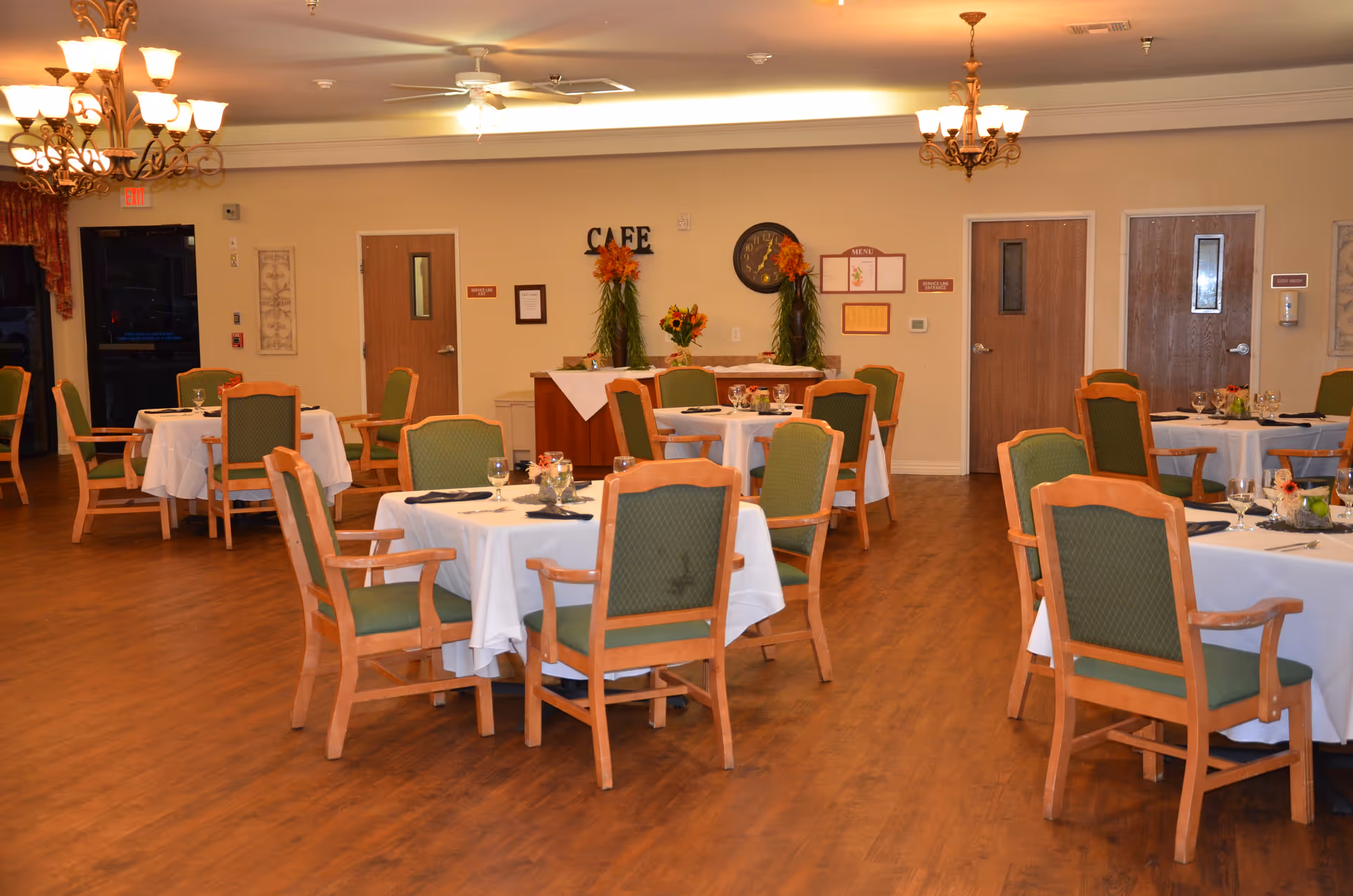 A dining room in a senior living facility with round tables covered in white tablecloths, each surrounded by wooden chairs with green cushions. The room has wooden flooring, chandeliers hanging from the ceiling, and a small buffet area at the back with floral decorations and a sign that reads 'CAFE'.