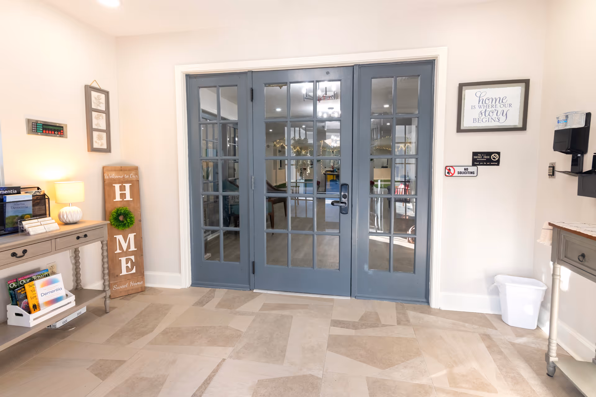 Interior view of a senior living facility entrance area with blue double glass doors. On the left side, there is a small table with a lamp, brochures, and a wooden sign that reads 'HOME' with a green wreath on the letter O. On the right side, there is a framed sign that says 'Home is where our story begins,' a hand sanitizer dispenser, and a small trash bin. The floor has a patterned tile design.
