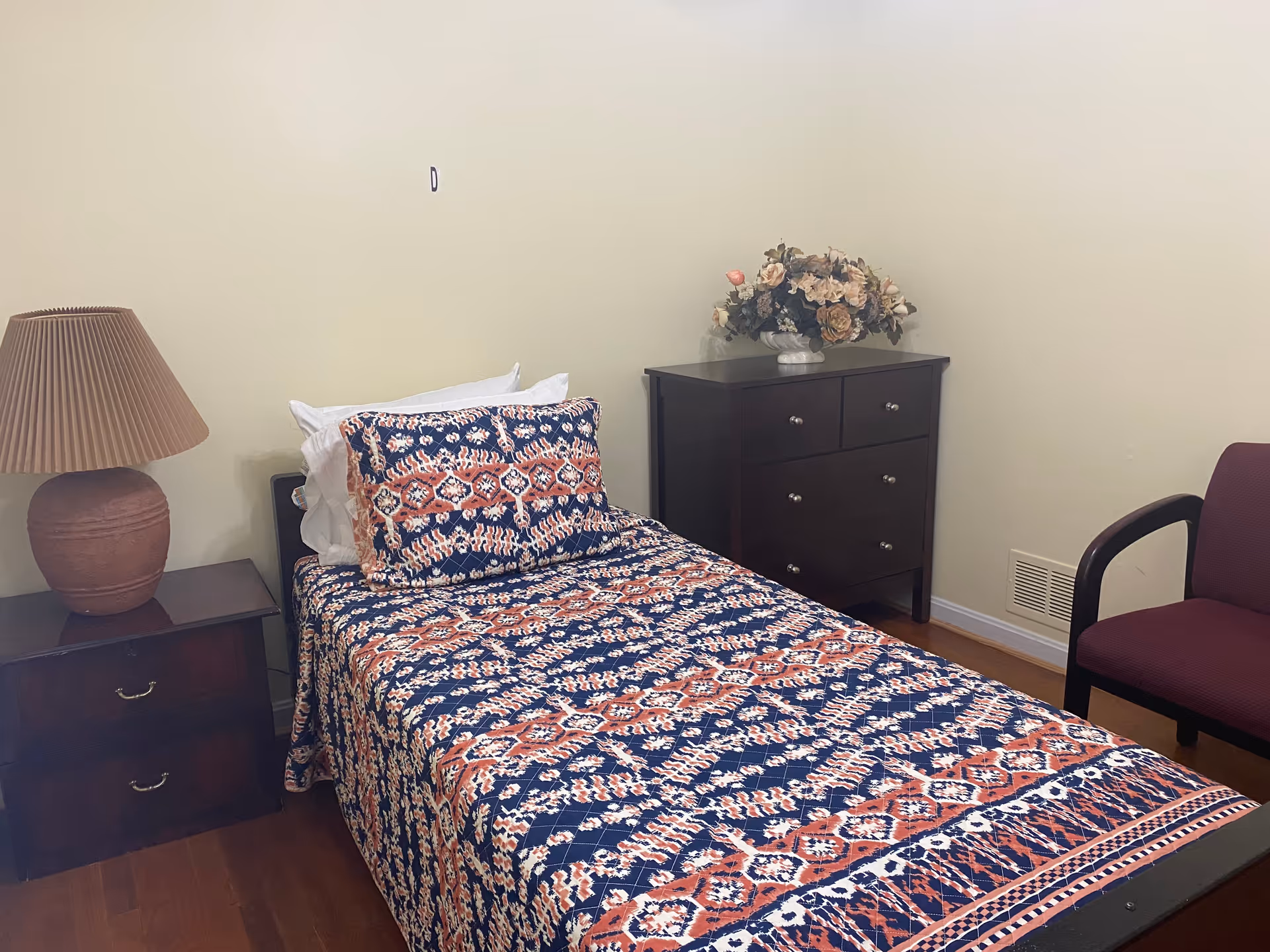 Bedroom scene with a patterned bedspread, dresser, nightstand, lamp, and flowers.