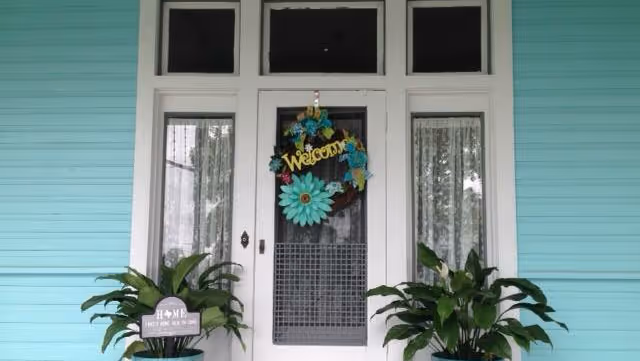 Turquoise building exterior with white-framed glass door, welcome wreath, and two large potted plants.