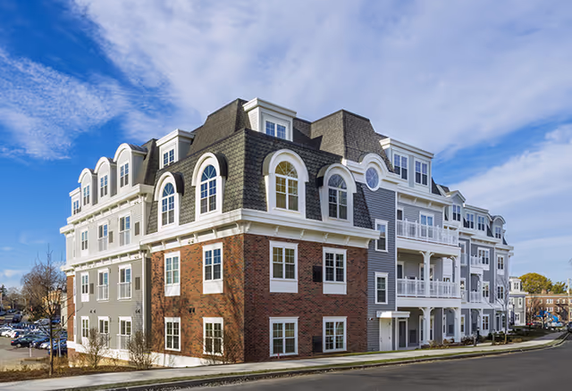 Exterior view of a multi-story senior living facility building with a combination of brick and gray siding, featuring arched windows and balconies under a partly cloudy blue sky.