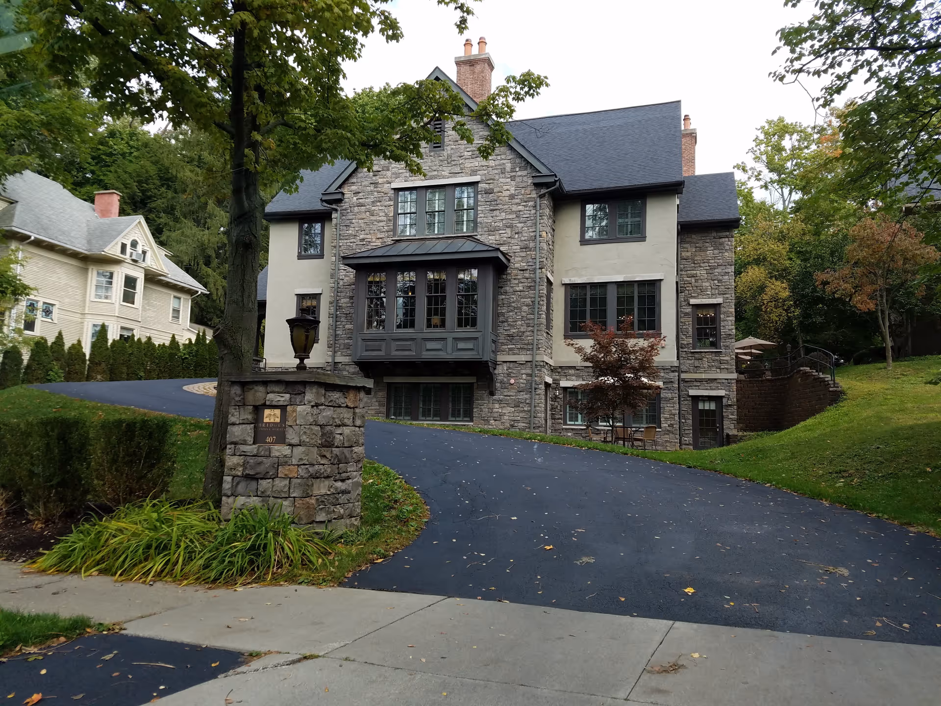 Tudor-style senior living building with stone facade, winding driveway, and surrounding mature trees.