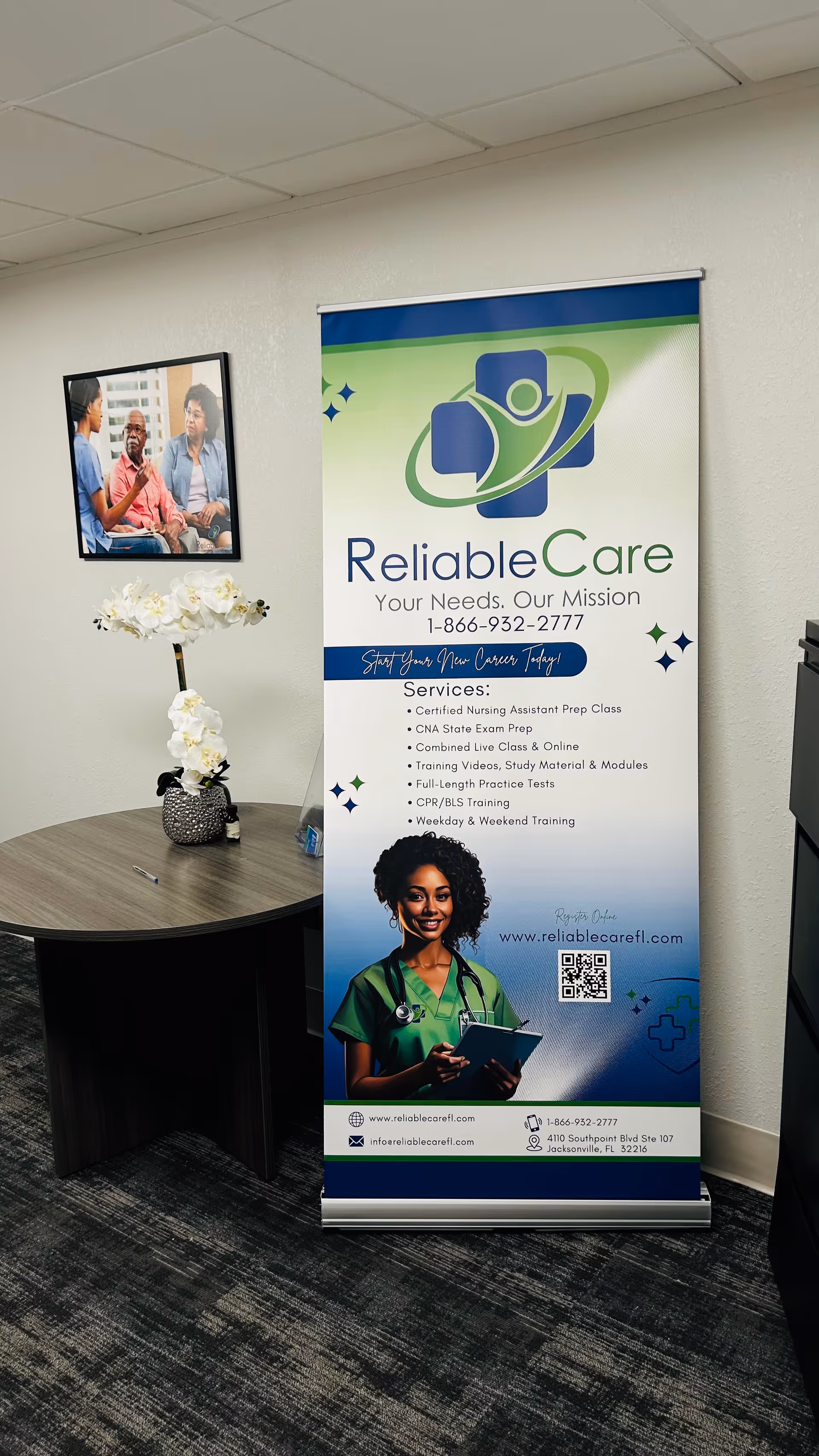 An interior corner with a Reliable Care roll-up banner, table, flowers, and framed photo.