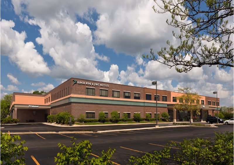 Exterior brick medical facility with a parking lot, manicured shrubs, and a blue sky.