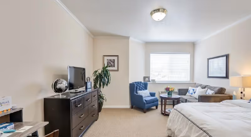 Bright hotel-style senior living suite with a bed, TV, dresser, and seating area.