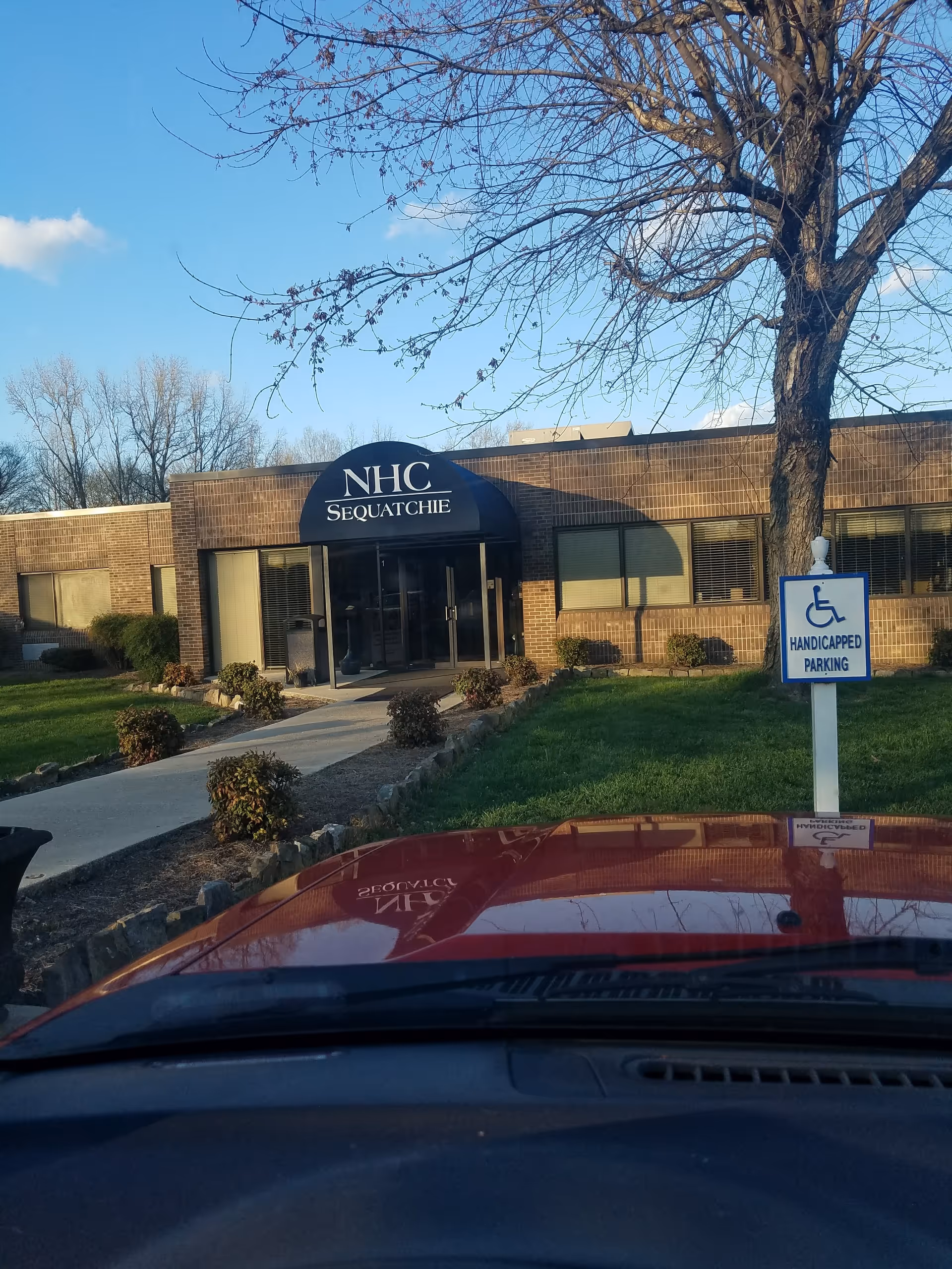 Exterior view of NHC Healthcare - Sequatchie entrance with blue awning, wheelchair sign, and parked car.