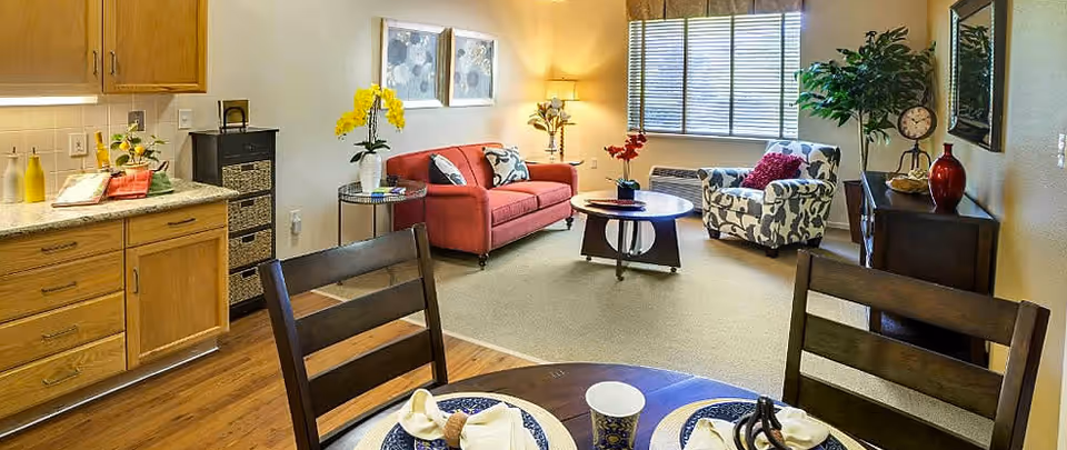 Open-plan living room and kitchenette with a red sofa, patterned armchair, dining table in the foreground, and a window letting in light.