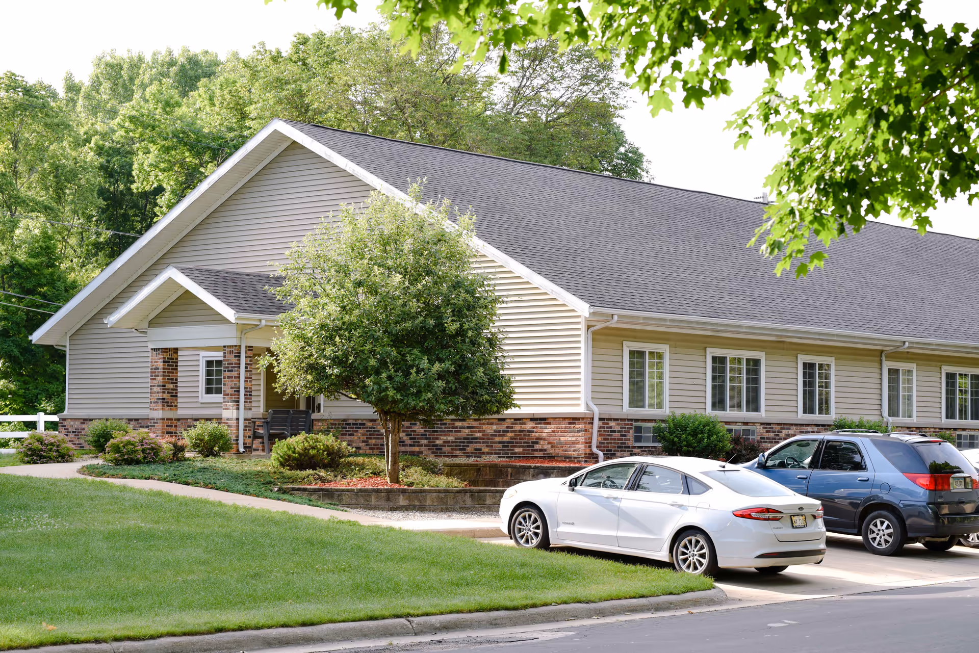 Exterior view of a single-story building with beige siding and brick accents, surrounded by green trees and shrubs. Two cars are parked in front of the building on a paved driveway. The scene is bright and sunny with a well-maintained lawn in the foreground.