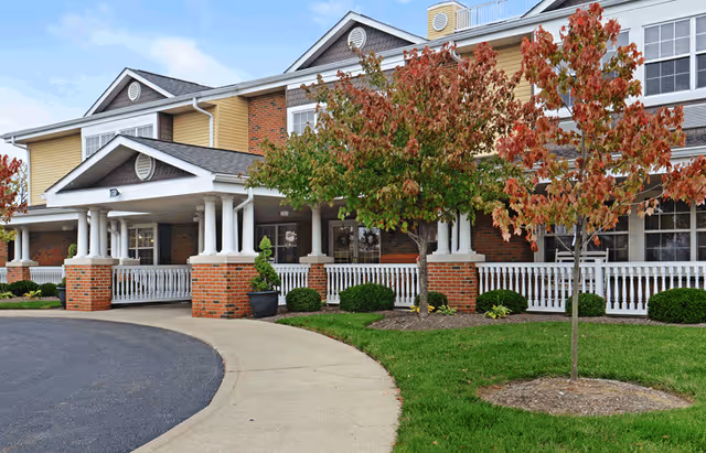 Exterior front of a senior living building with a covered porch, white columns, brick facade, walkway and landscaped lawn with small trees.