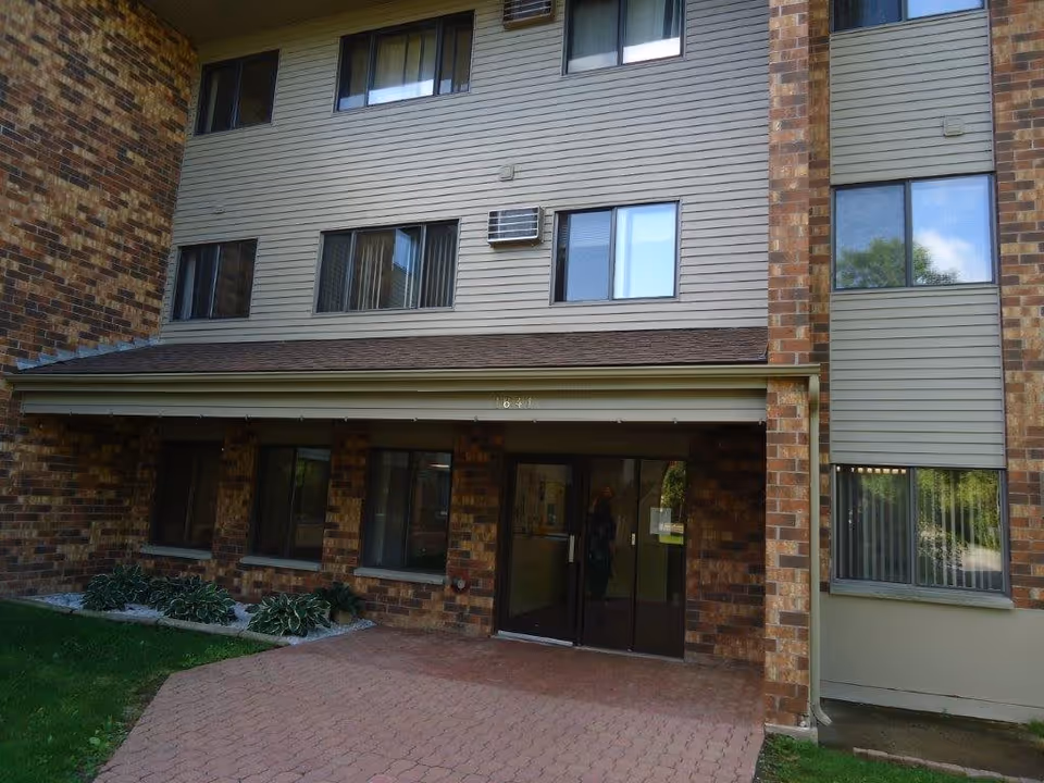 Exterior view of a senior living facility entrance with brick and siding walls, multiple windows, a glass door entrance, and a small garden area with plants beside the walkway.