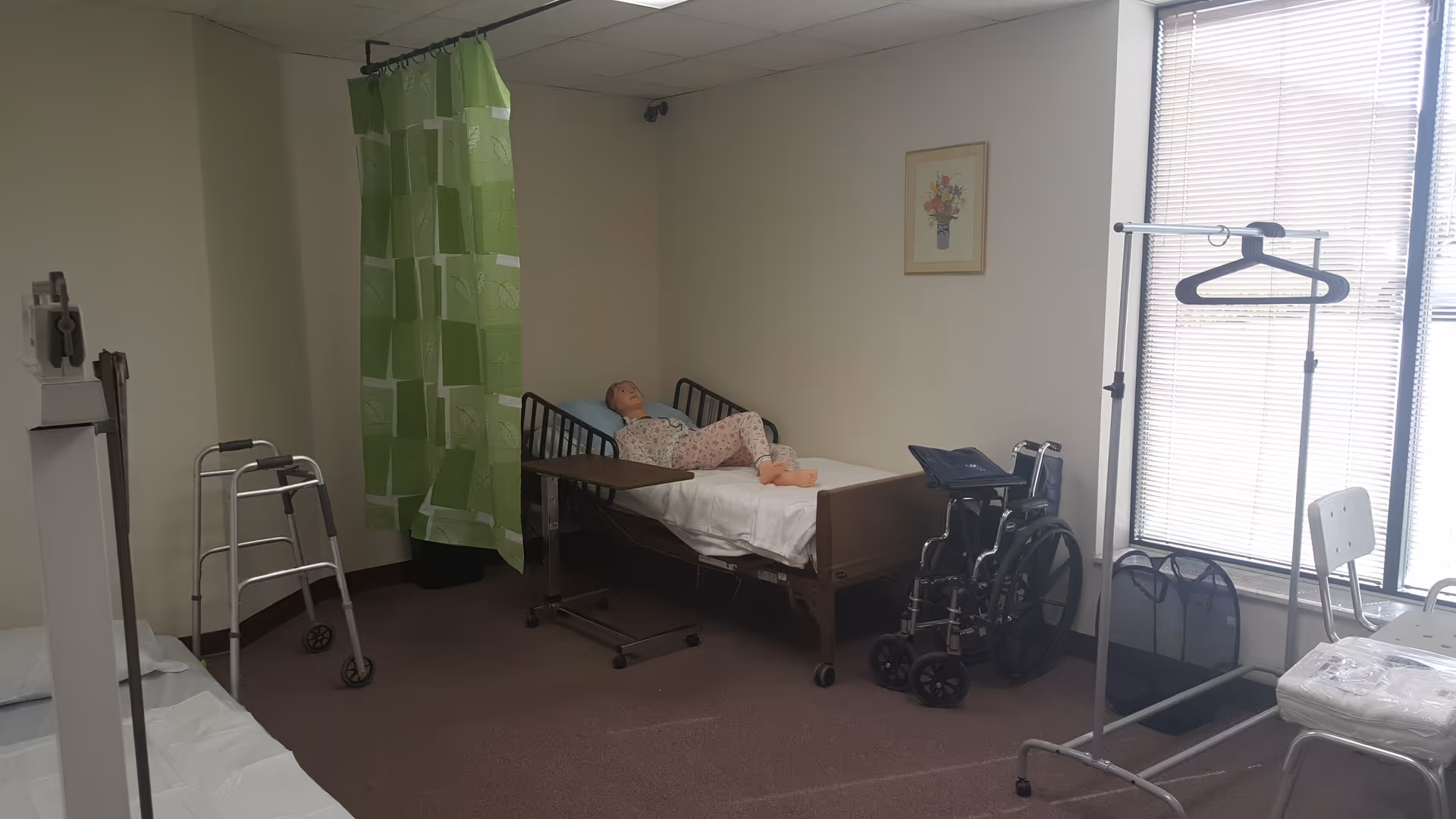Hospital-style patient room with bed, wheelchair, green privacy curtain, and medical equipment.