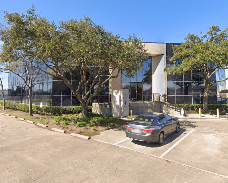 Glass-front building with trees, a parking lot, and a car in front.