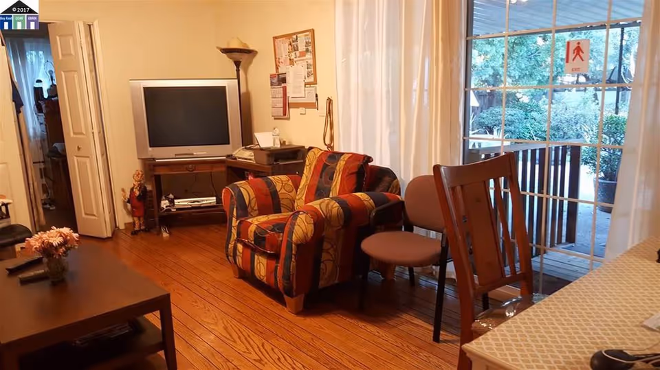 Cozy living room with a patterned armchair, TV on a stand, coffee table, extra chairs, and sliding glass doors leading to an outdoor porch.