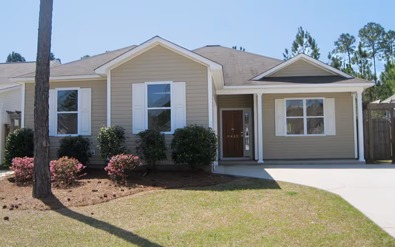 Single-story beige house with front yard, shrubs, and a driveway.