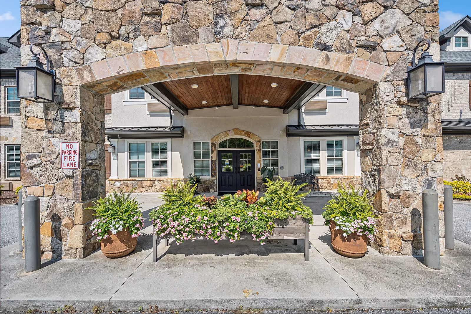 Stone arched entrance under wooden canopy, planters and bench in front of a senior living building.