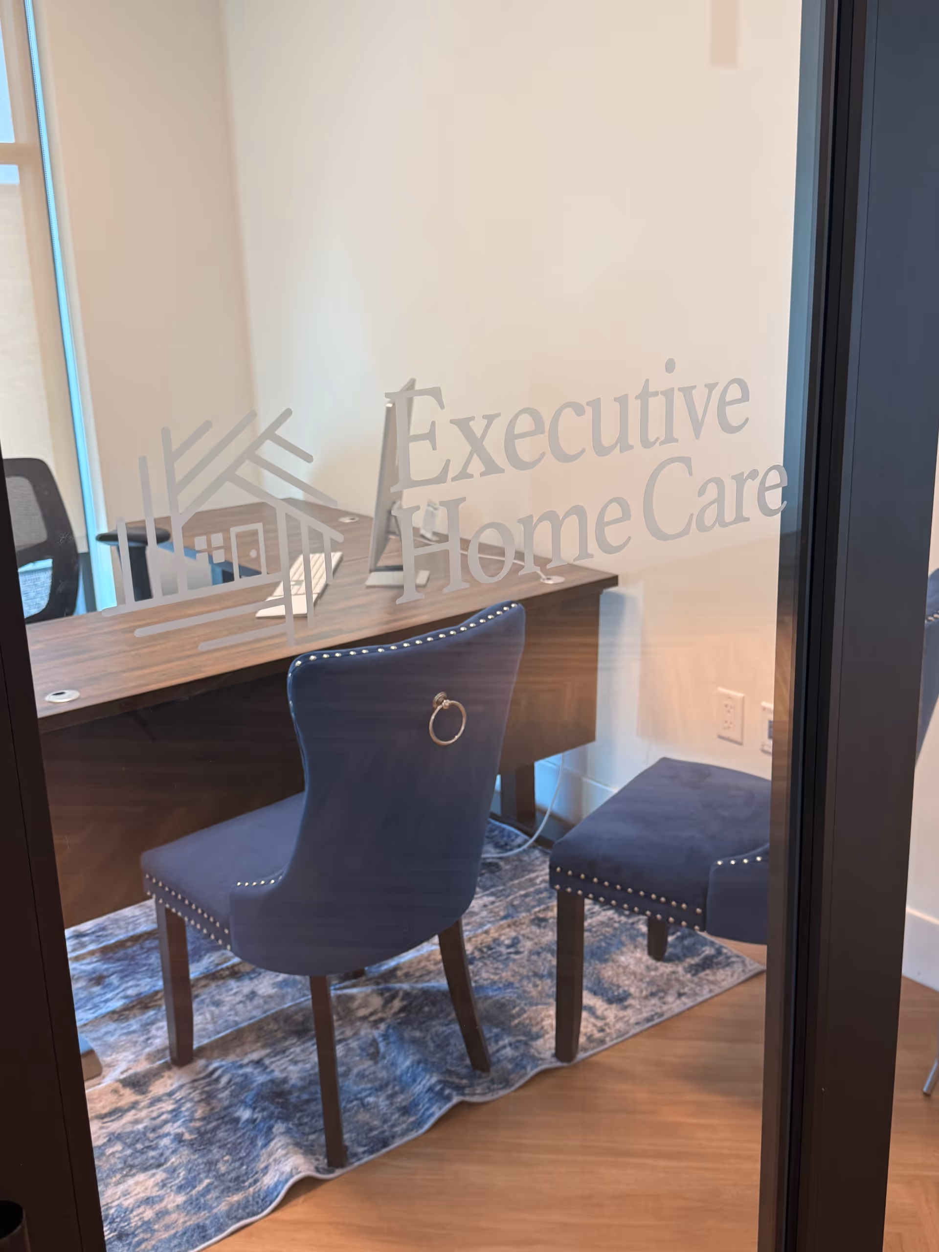 Office workspace with desk, blue chair, and glass door showing Executive Home Care branding.