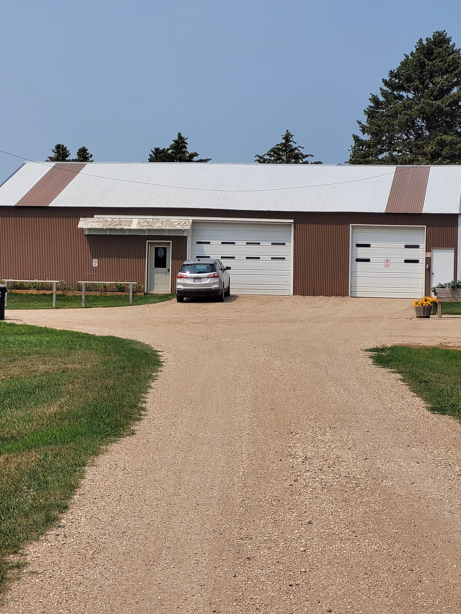 Gravel driveway leads to a brown metal building with two large garage doors and a car.