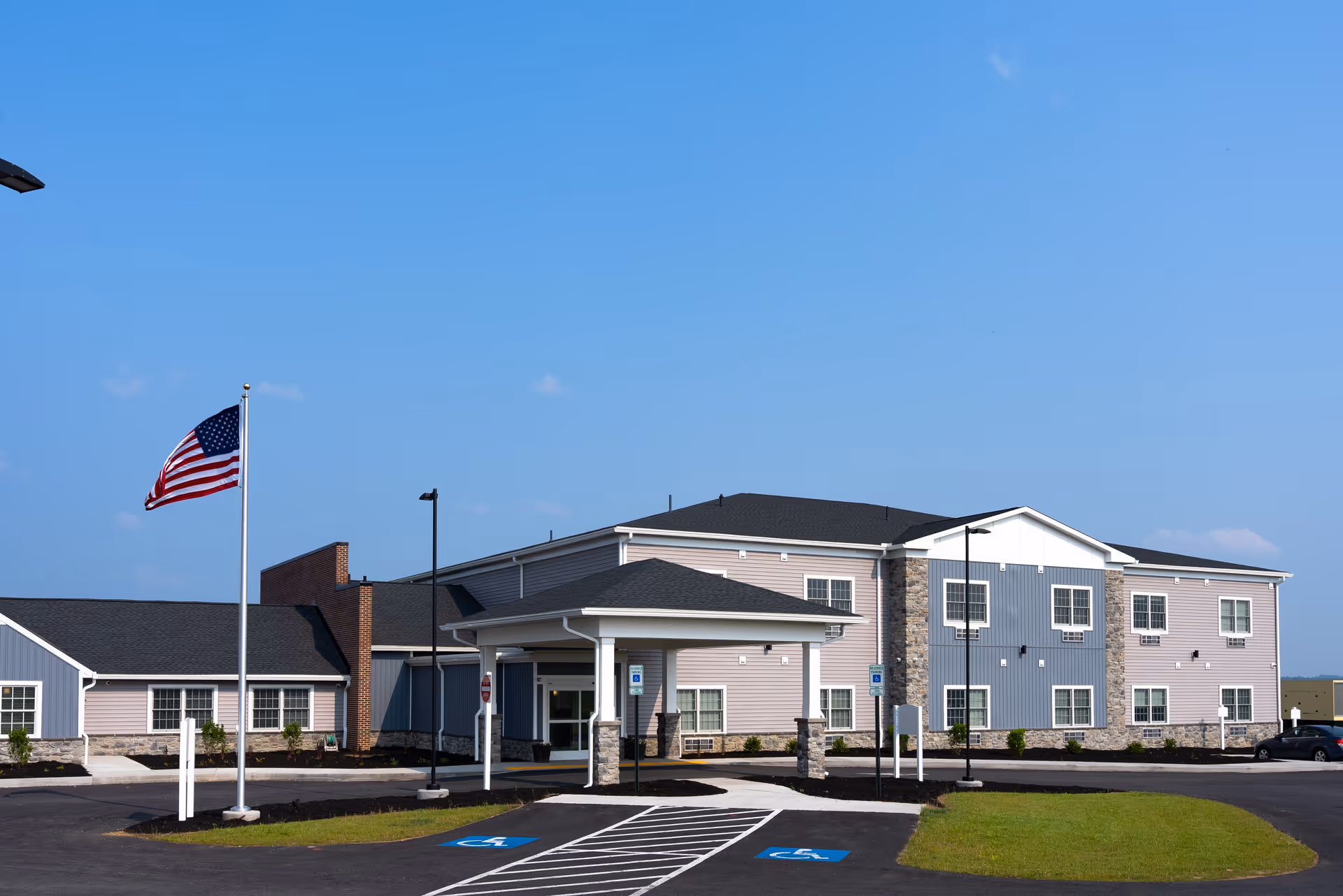 Exterior view of Heritage Hills Retirement Home building under a clear blue sky, featuring a two-story structure with a covered entrance, an American flag on a flagpole, and a parking area with handicapped spaces in front.