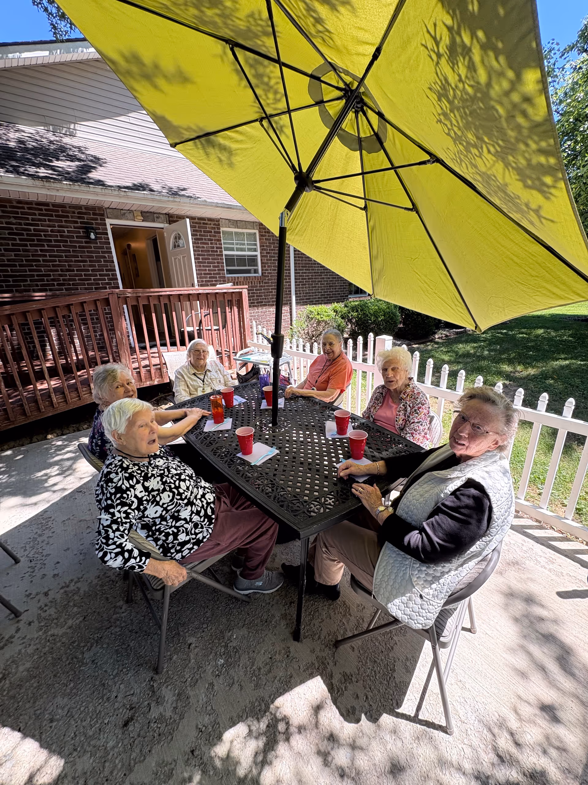Six elderly residents sit around a metal table under a large yellow umbrella outdoors.