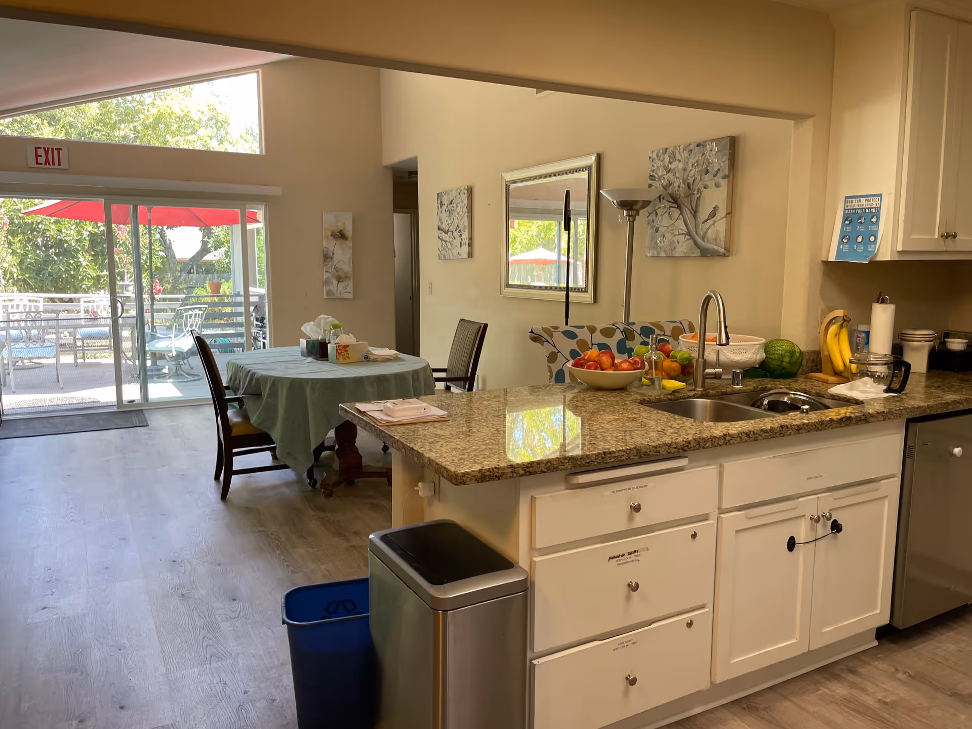 Interior view of a kitchen and dining area in a senior living facility. The kitchen features a granite countertop with a sink, a bowl of fruit, bananas, a watermelon, and kitchen appliances. Adjacent to the kitchen is a dining table covered with a green tablecloth and surrounded by chairs. Large sliding glass doors lead to an outdoor patio with red umbrellas and seating. The room is well-lit with natural light and decorated with wall art and a mirror.