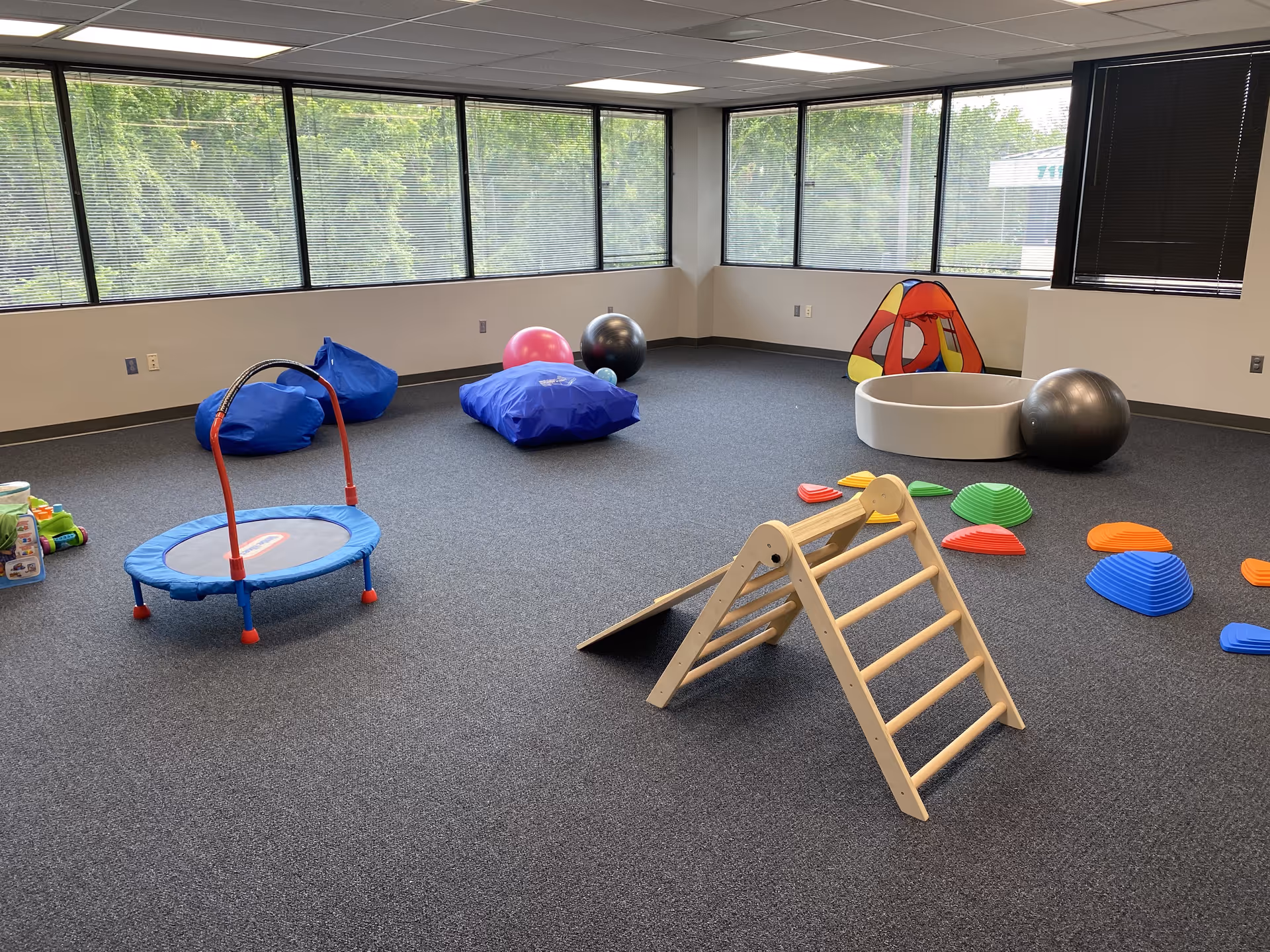 Indoor playroom with climbing structure, trampoline, soft mats, tunnel tent, and windows.