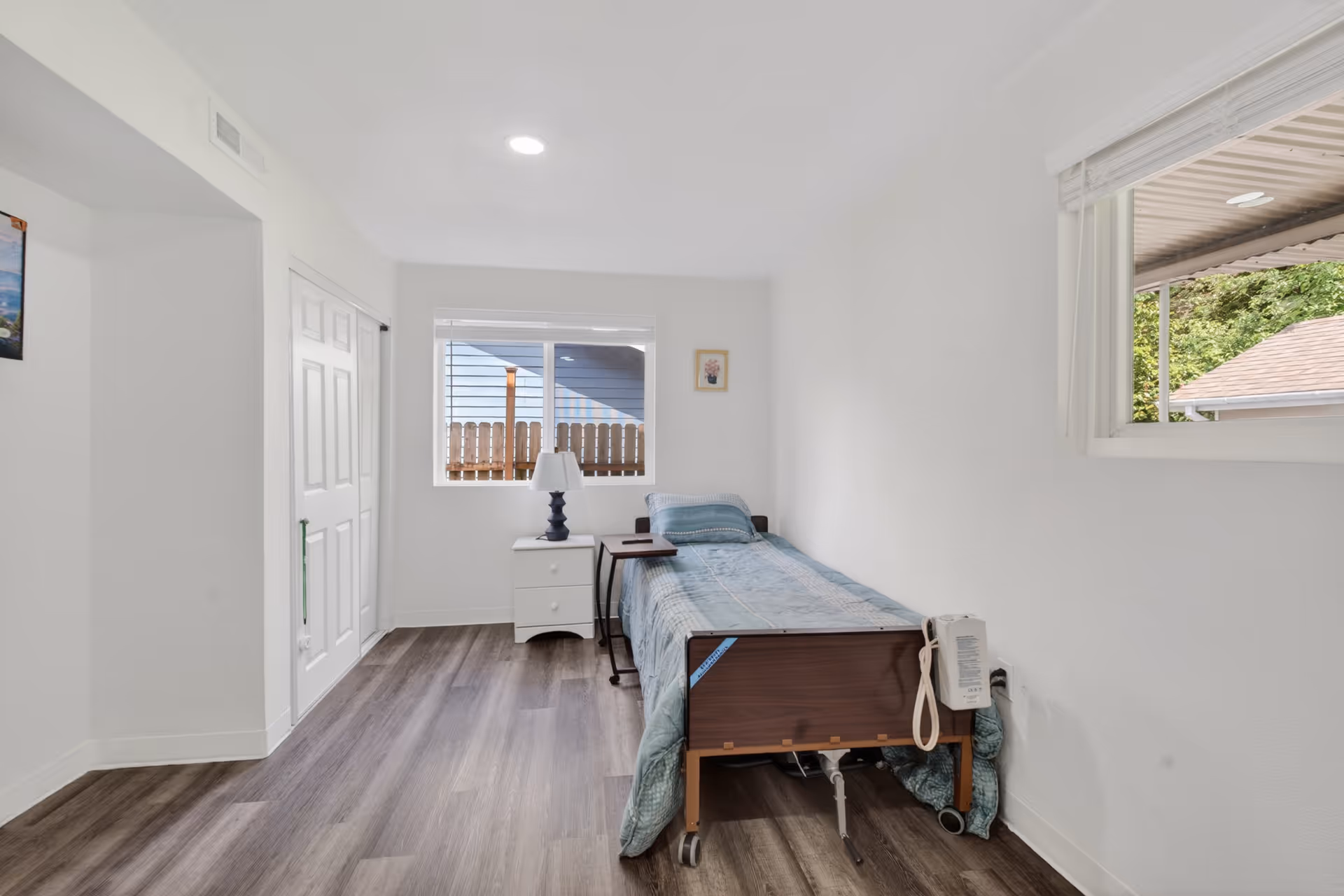 A small, bright bedroom with a single bed, nightstand, lamp, and windows.