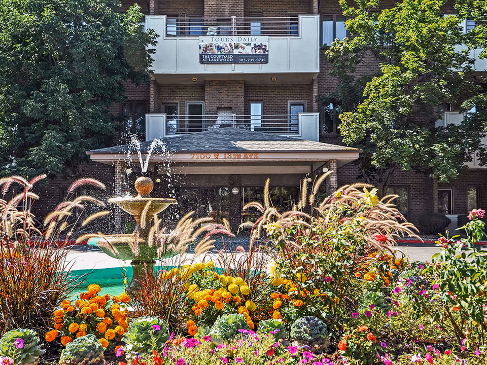 Entrance of The Courtyard at Lakewood senior living building with a fountain and colorful flowerbeds in the foreground.