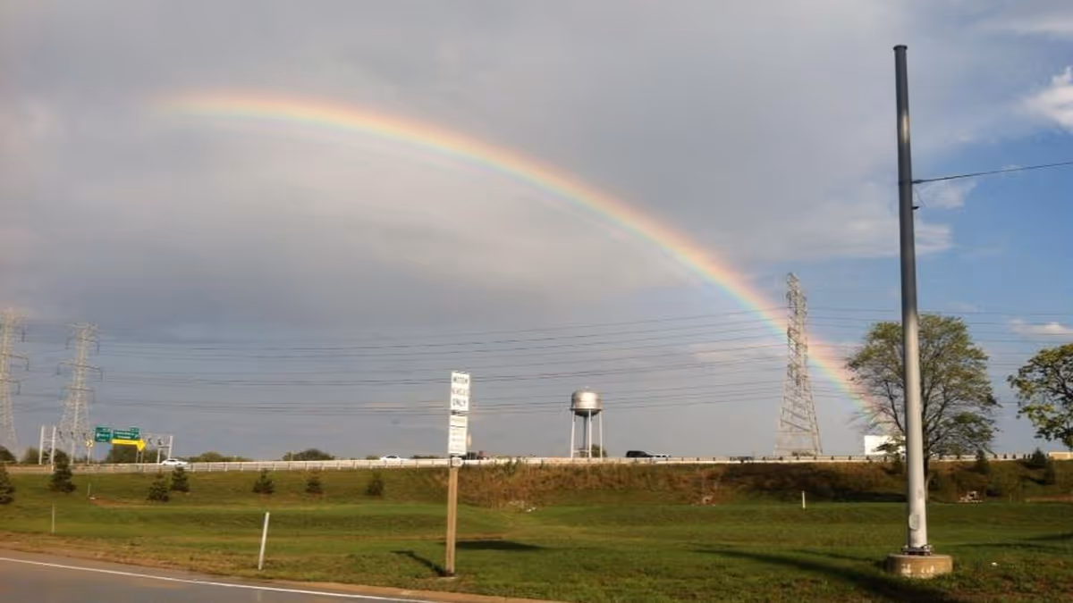 A rainbow arches over a grassy highway scene with power lines and trees.