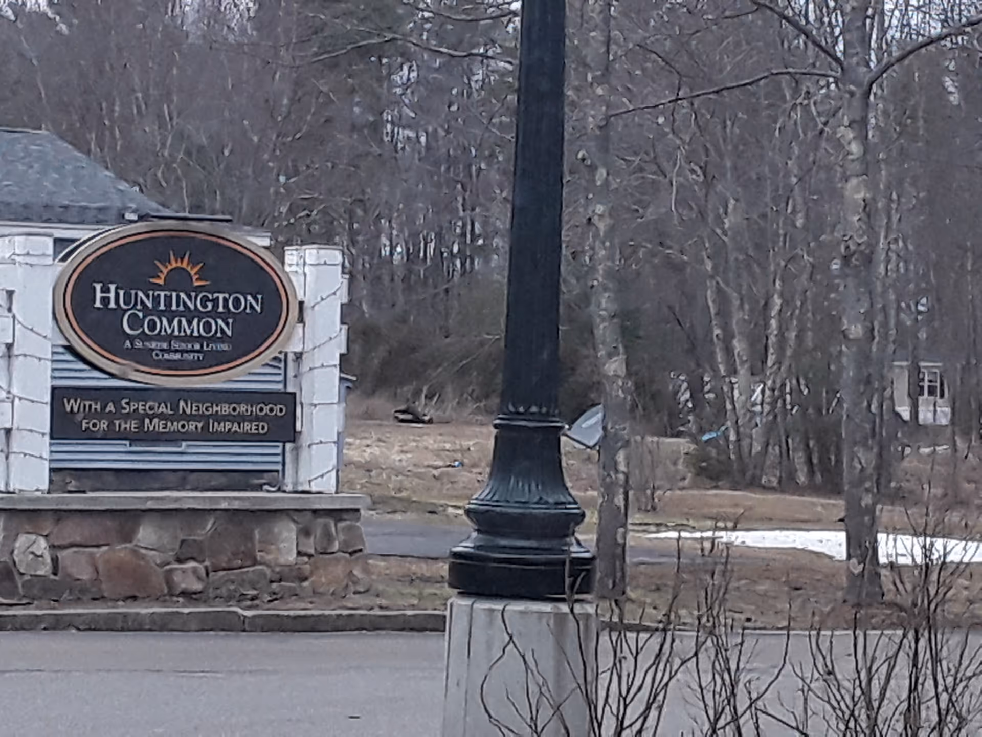 Outdoor entrance sign reading Huntington Common sits beside a lamp post amid bare trees.