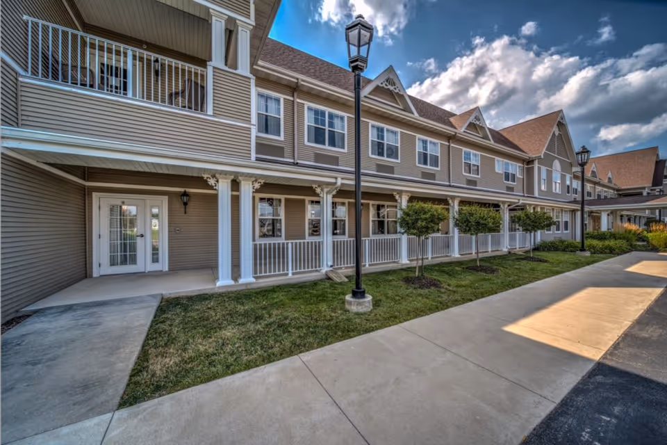 Two-story senior living building exterior with a covered porch, white railings, lamp posts, walkway and manicured lawn under a partly cloudy sky.