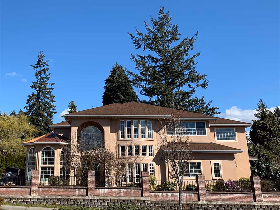 Two-story beige senior living facility with brick fence and tall trees against a blue sky.