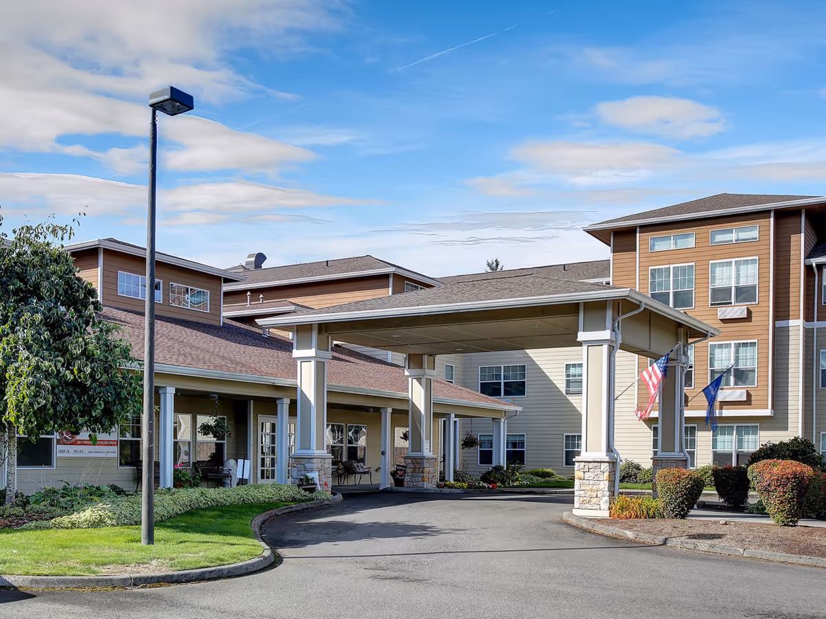 Covered entrance and driveway of a multi-story senior living building with flags and landscaped grounds.
