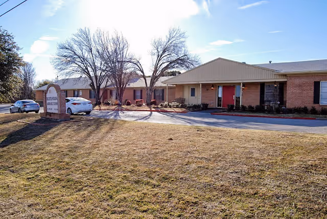 Exterior view of a single-story brick building with a beige roof and red doors, surrounded by a lawn and leafless trees under a clear blue sky. Two cars are parked near a sign that reads 'Heritage Place of Decatur'.