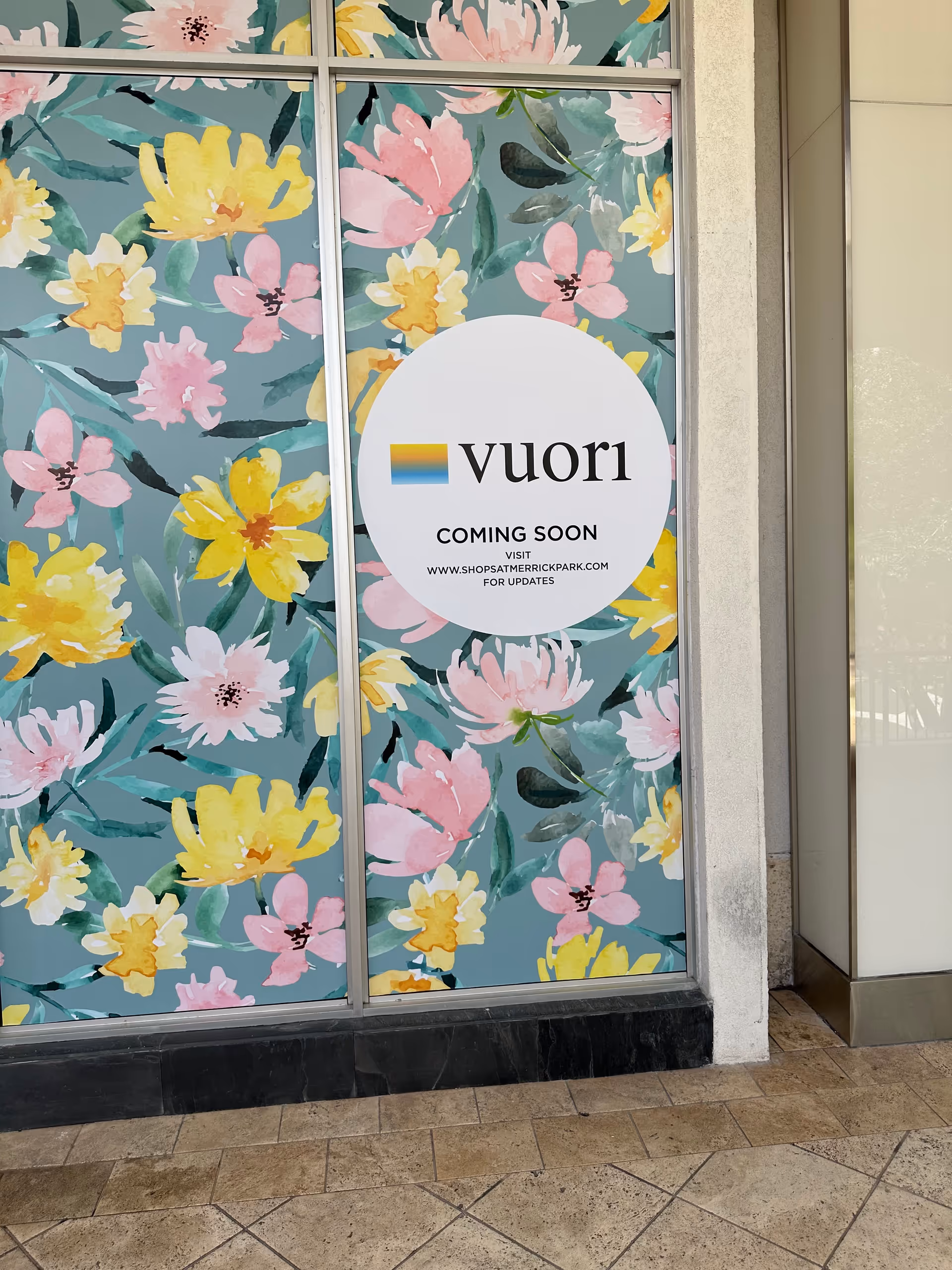 Glass storefront window with colorful floral panels and a circular Vuori Coming Soon sign.
