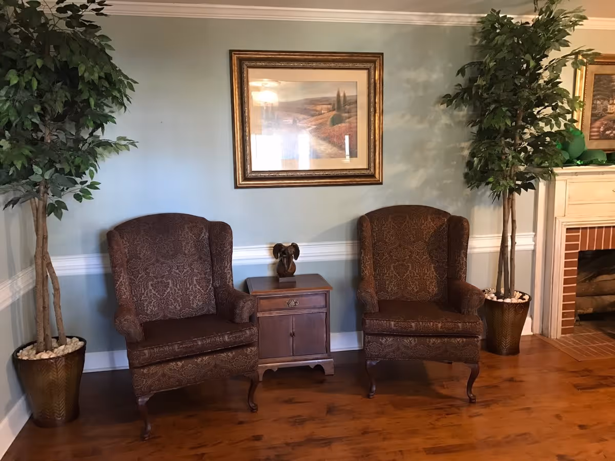 Two upholstered armchairs flanking a small wooden side table beneath a framed painting in a cozy living room with potted trees and a fireplace.
