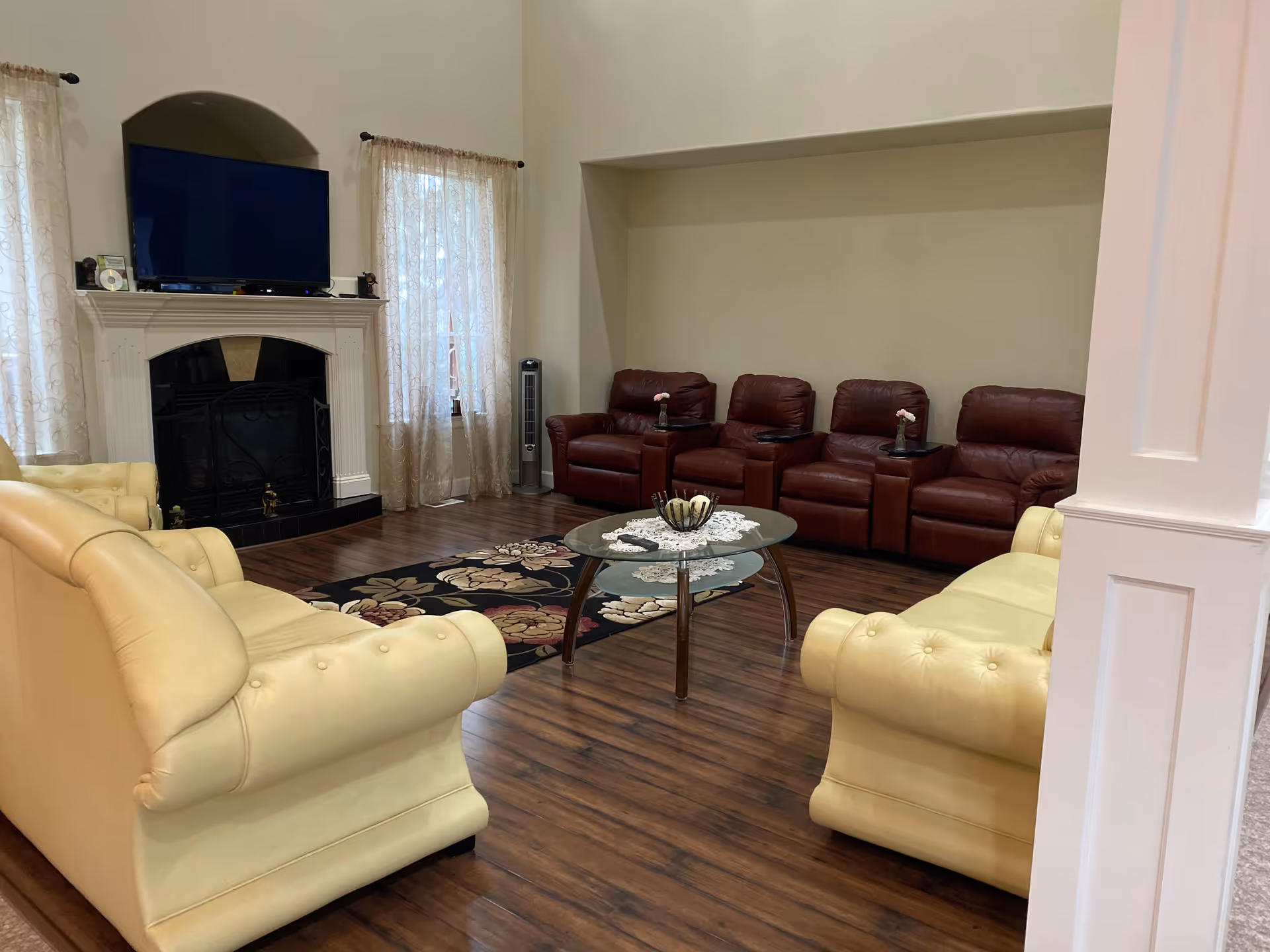 Spacious living room with yellow sofas facing a glass coffee table, a fireplace with a TV above, and a row of brown recliners along the far wall.