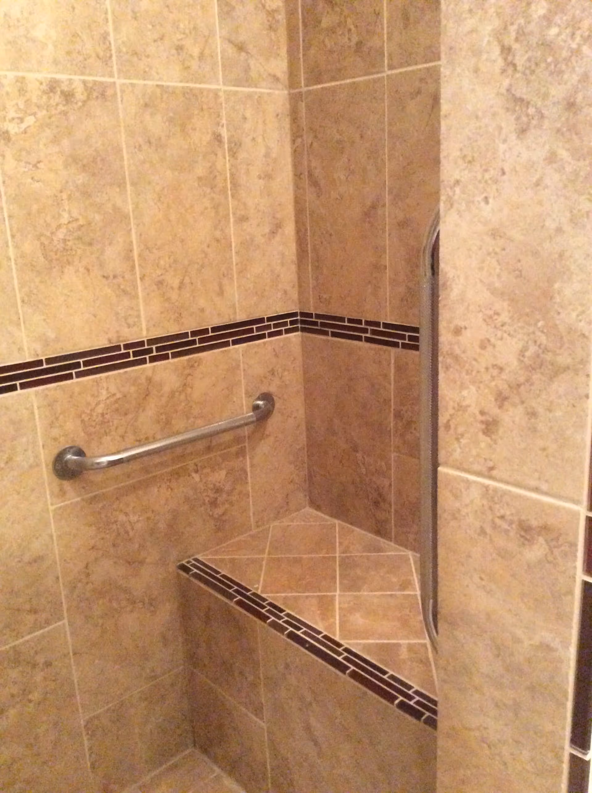 Corner of a tiled shower with beige and brown tiles, featuring a built-in triangular bench and a horizontal metal grab bar on the wall.