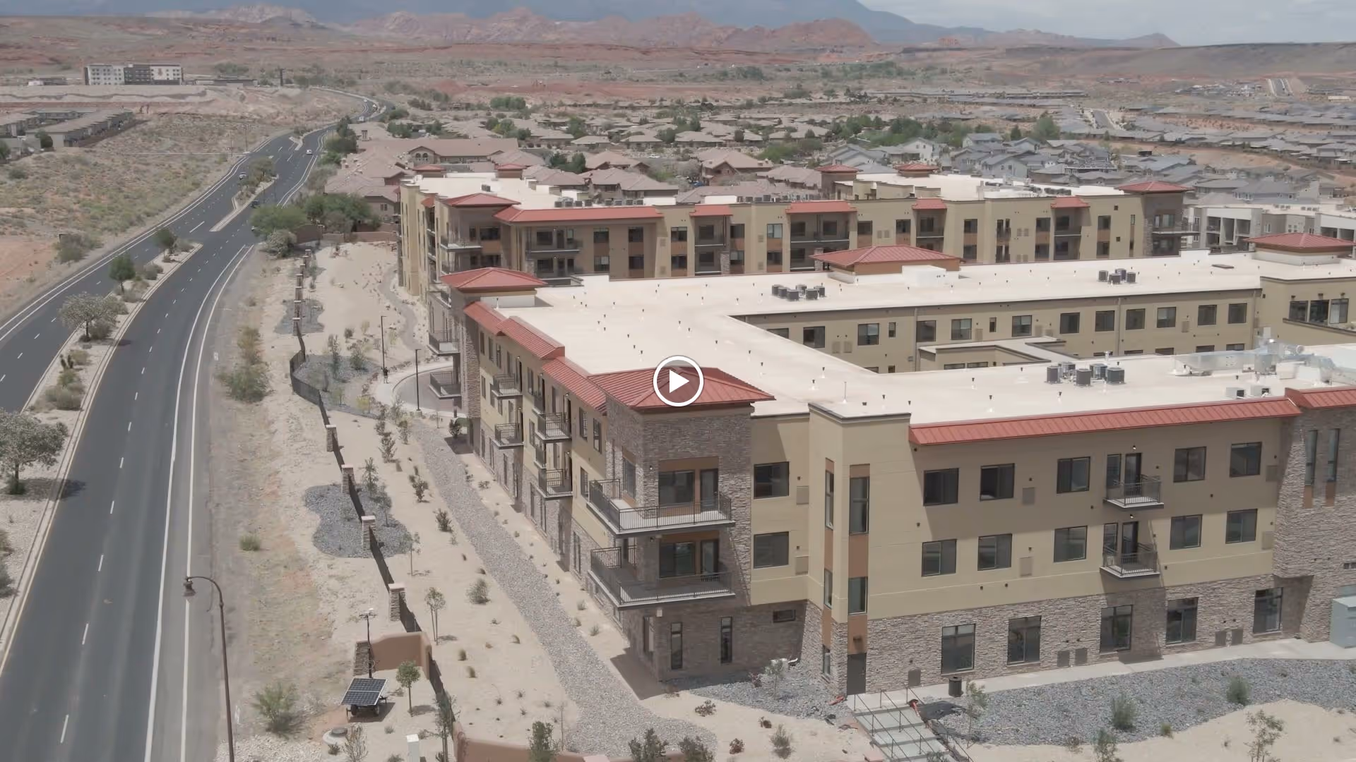 Aerial view of a large, modern senior living facility with beige and stone exterior walls and red roofs, situated in a desert landscape with mountains in the background and a road running alongside the building.