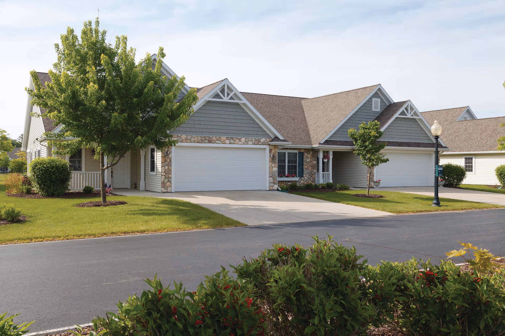 Row of single-story residential units with attached garages, manicured lawns, trees, and a street in front.