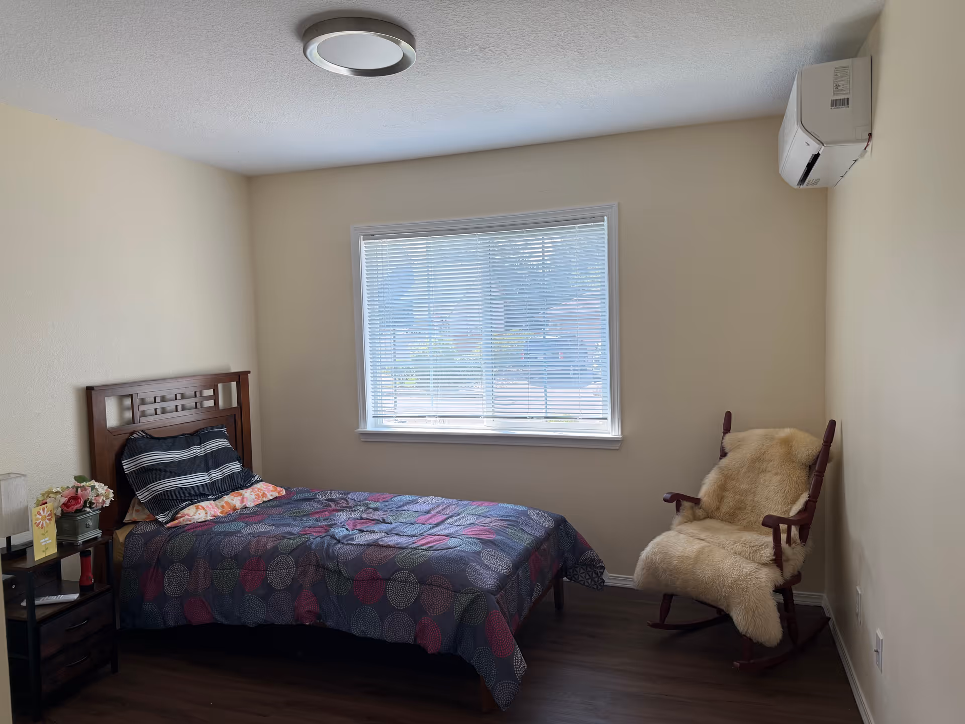 A small bedroom with a bed, nightstand, rocking chair, window, and an air conditioner.