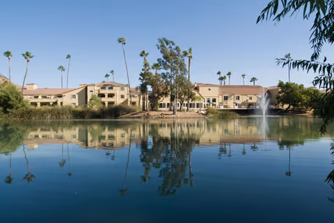 View of a senior living facility building named Savant of Jurupa Valley reflected in a calm pond with a water fountain, surrounded by palm trees and clear blue sky.
