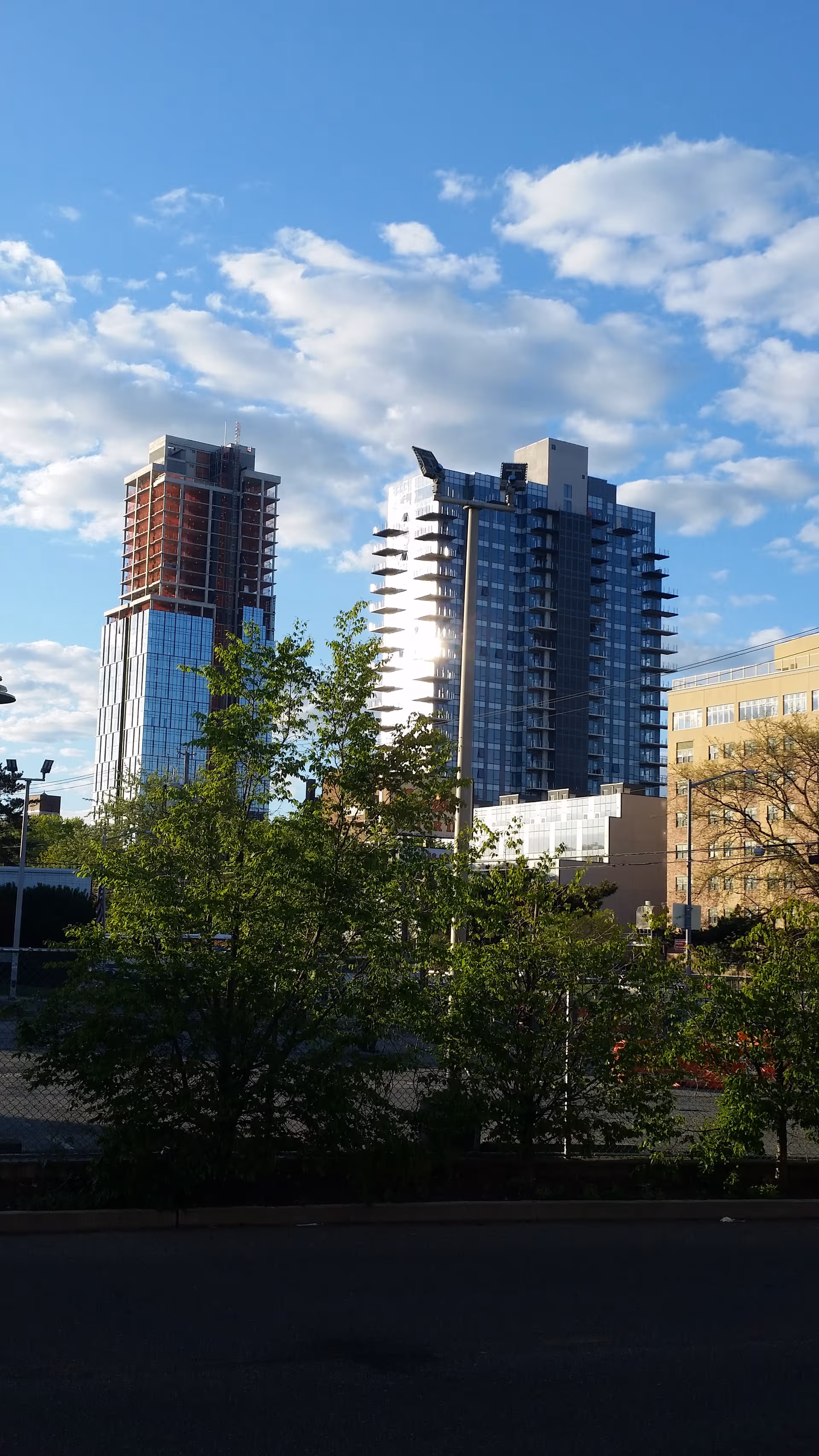 Tall modern skyscrapers rise above green trees under a blue sky with clouds.