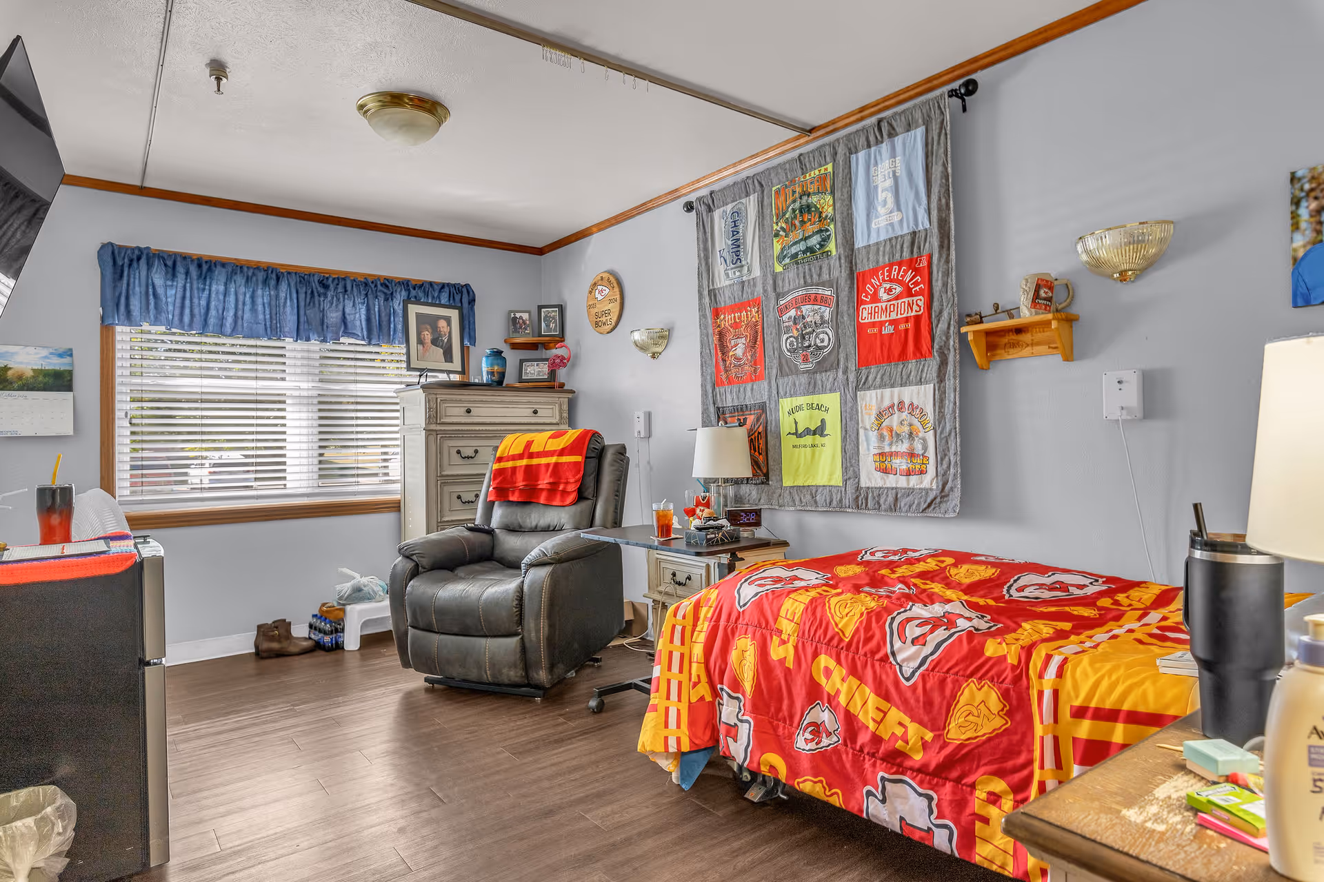 A cozy room in a senior living facility featuring a single bed with a red and yellow Kansas City Chiefs blanket, a gray recliner chair with a red and yellow blanket draped over it, a wooden dresser, a side table with a lamp and drinks, and a large window with blue curtains. The walls are decorated with framed photos, a quilt with various patches, and a small wooden shelf.