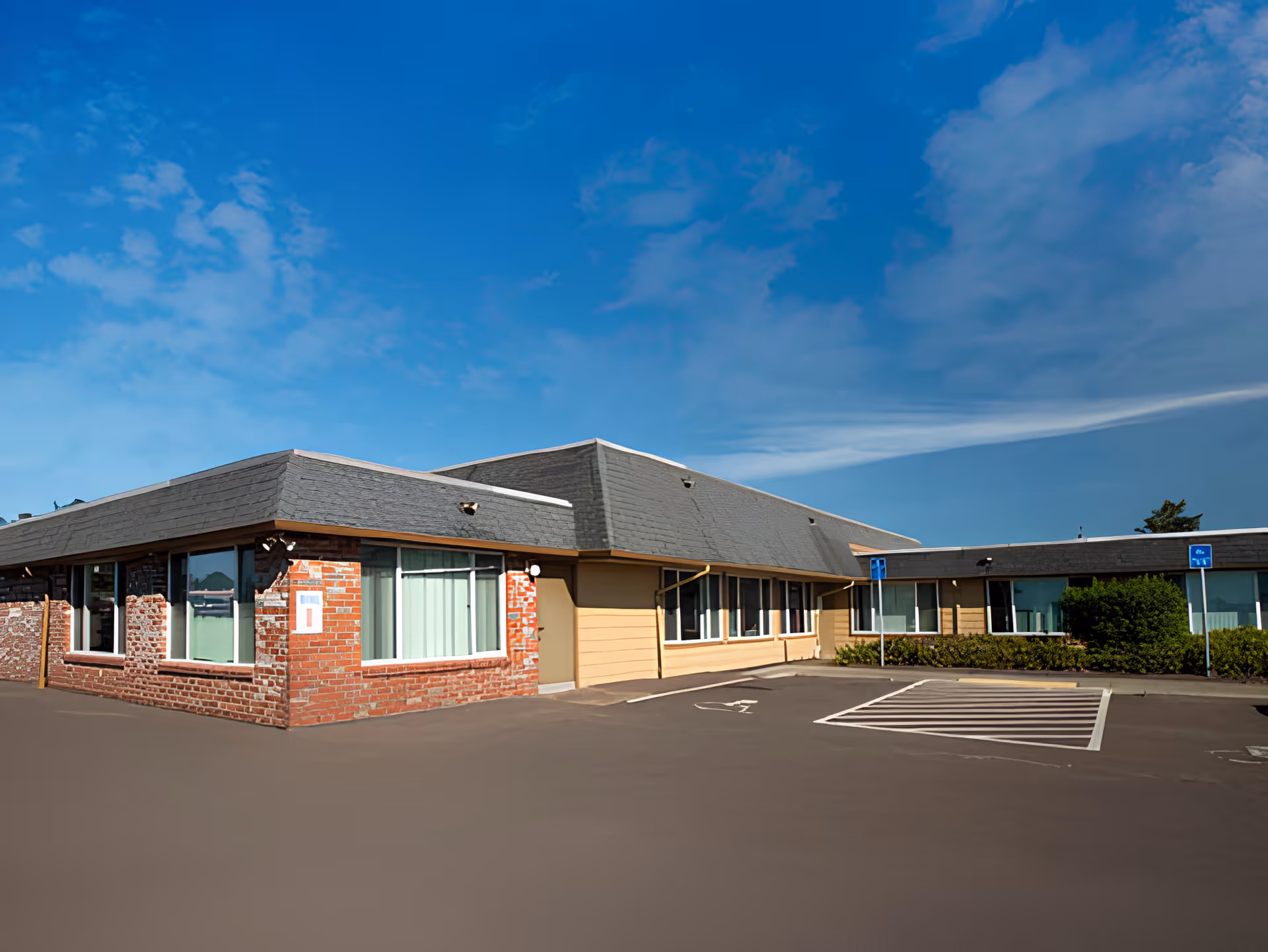 Exterior view of a single-story brick and siding building under a blue sky with some clouds. The building has multiple windows with curtains and a parking lot in front with marked handicap parking spaces.
