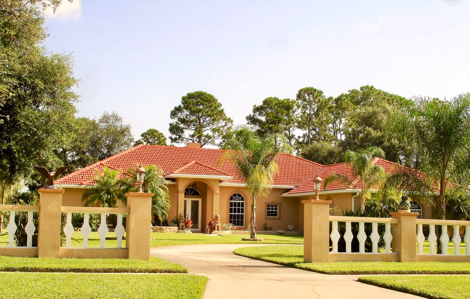 A single-story house with a red tiled roof surrounded by a beige fence with decorative pillars and lanterns. The house is set in a lush green yard with palm trees and other greenery, under a clear blue sky.