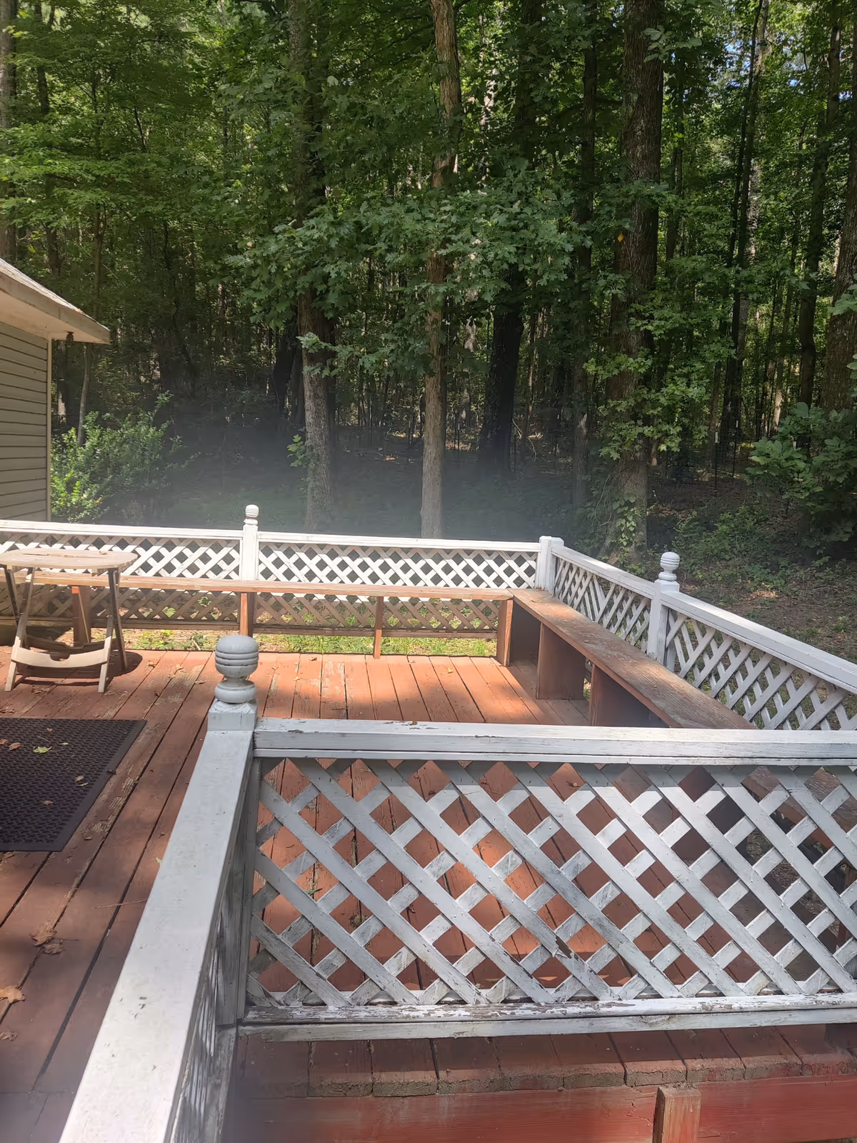 A wooden deck with white lattice railings and a built-in bench overlooking a dense wooded backyard.