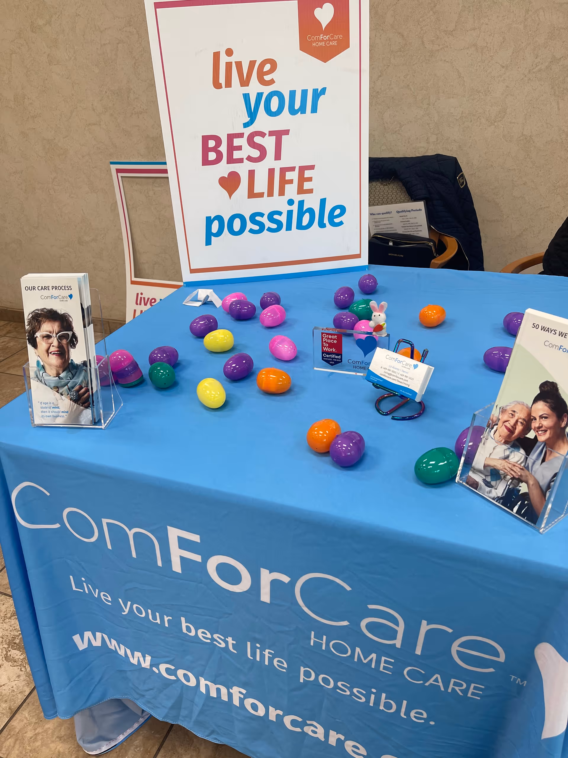 Blue ComForCare table with colorful eggs, brochures, and a sign indoors.