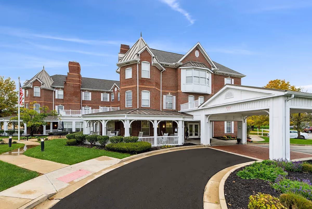 Exterior view of a large brick senior living facility with white trim and a covered entrance driveway. The building has multiple windows, a turret-like structure, and well-maintained landscaping with green bushes and trees. The sky is clear and blue.