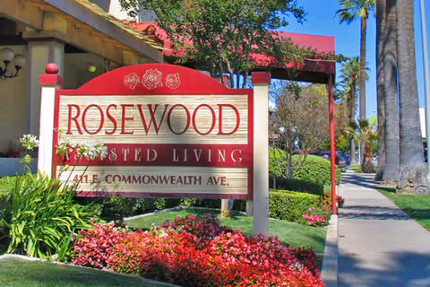 A red-and-cream wooden sign reading "ROSEWOOD Assisted Living 411 E. Commonwealth Ave." in front of landscaped beds, palm trees, and a sidewalk.