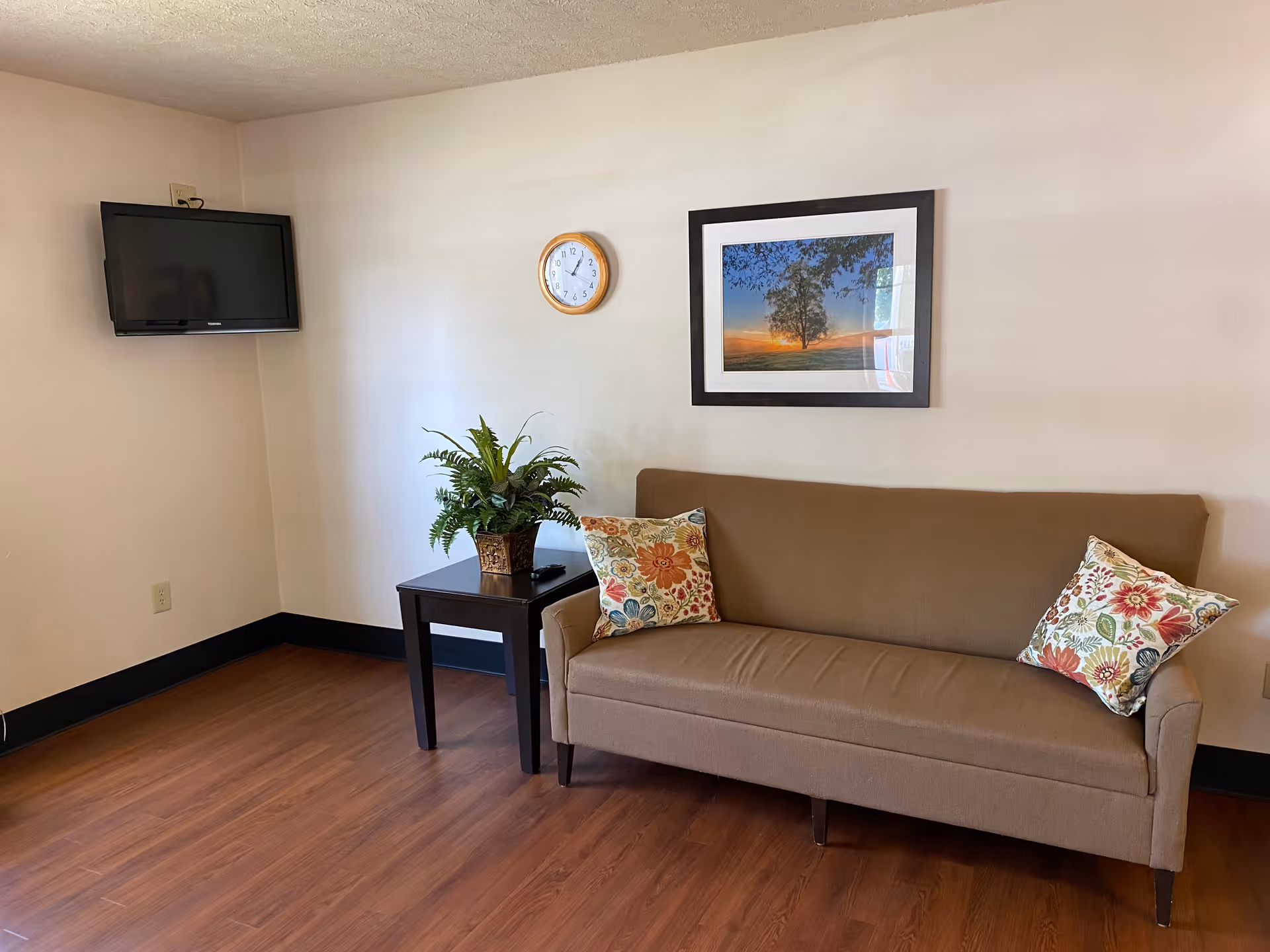 Neutral living area with brown sofa, side table, plant, clock, framed art, and mounted TV.